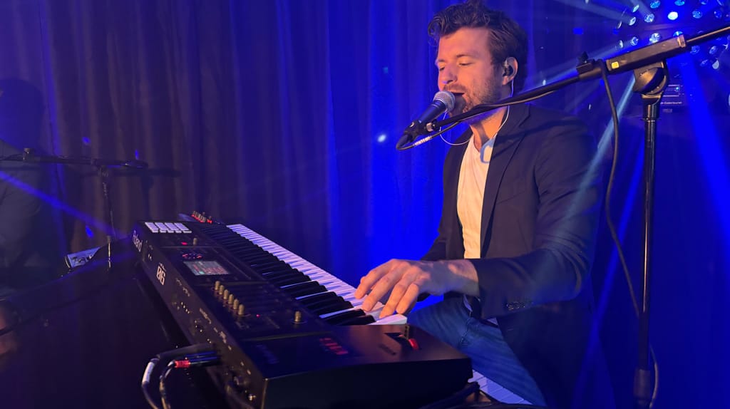 De pianist zingt achter zijn keyboard in blauw podiumlicht tijdens een optreden